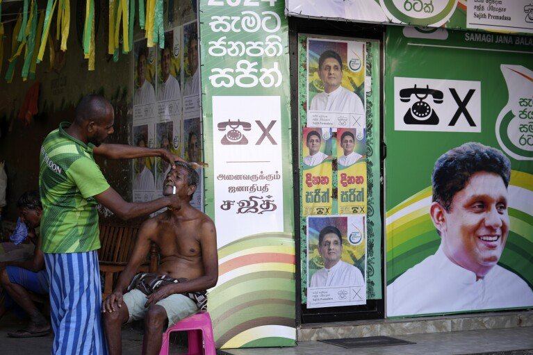 A man sits next to election posters of opposition leader Sajith Premadasa as he gets a shave from a roadside barber in Colombo, Sri Lanka, Tuesday, Sept. 17, 2024. (AP Photo/Rajesh Kumar Singh)